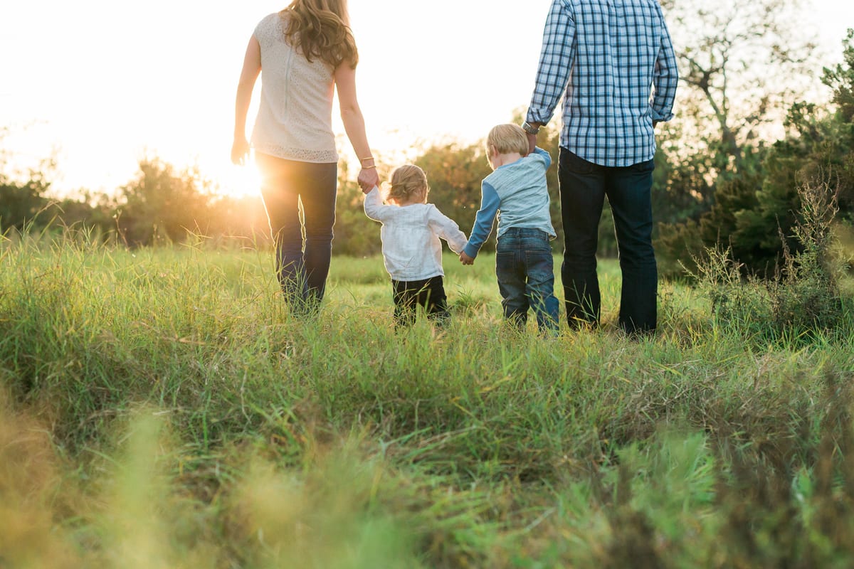 Parents walking hand in hand with their two young sons through a sunlit field