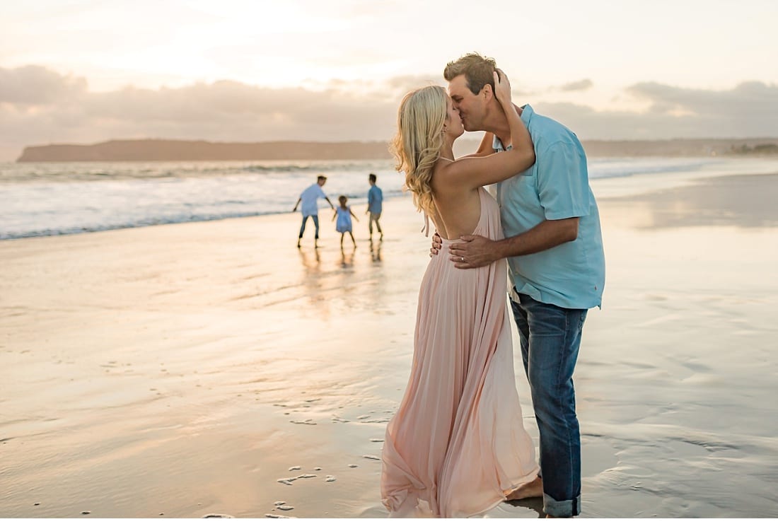 Couple kissing on Coronado Beach at sunset while their children play at the water's edge