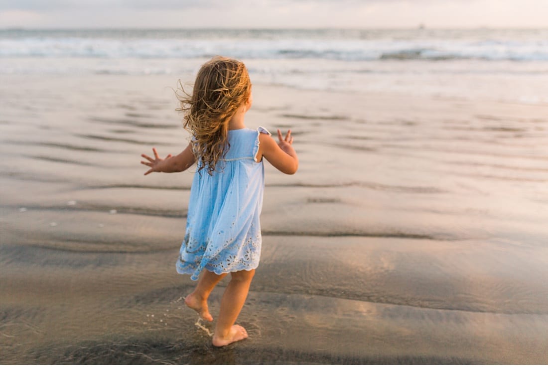 Little girl in a blue dress running barefoot toward the ocean waves