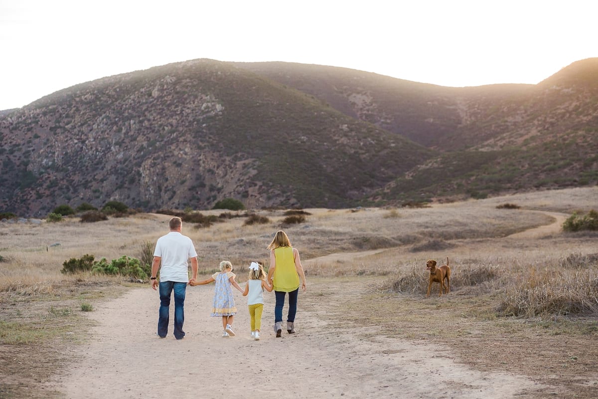 Family of four walking down a dirt trail toward the mountains with their dog