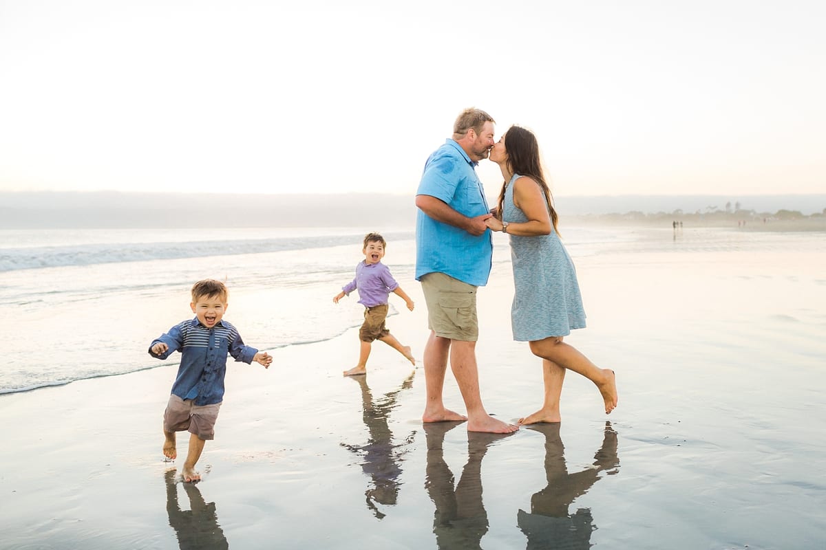 Parents kissing on the shore as their two boys run in the shallow water