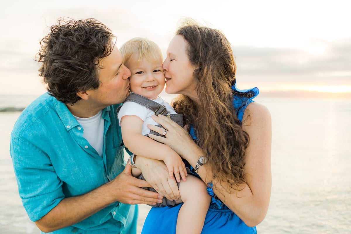 Parents kissing their smiling toddler son on both cheeks at the beach