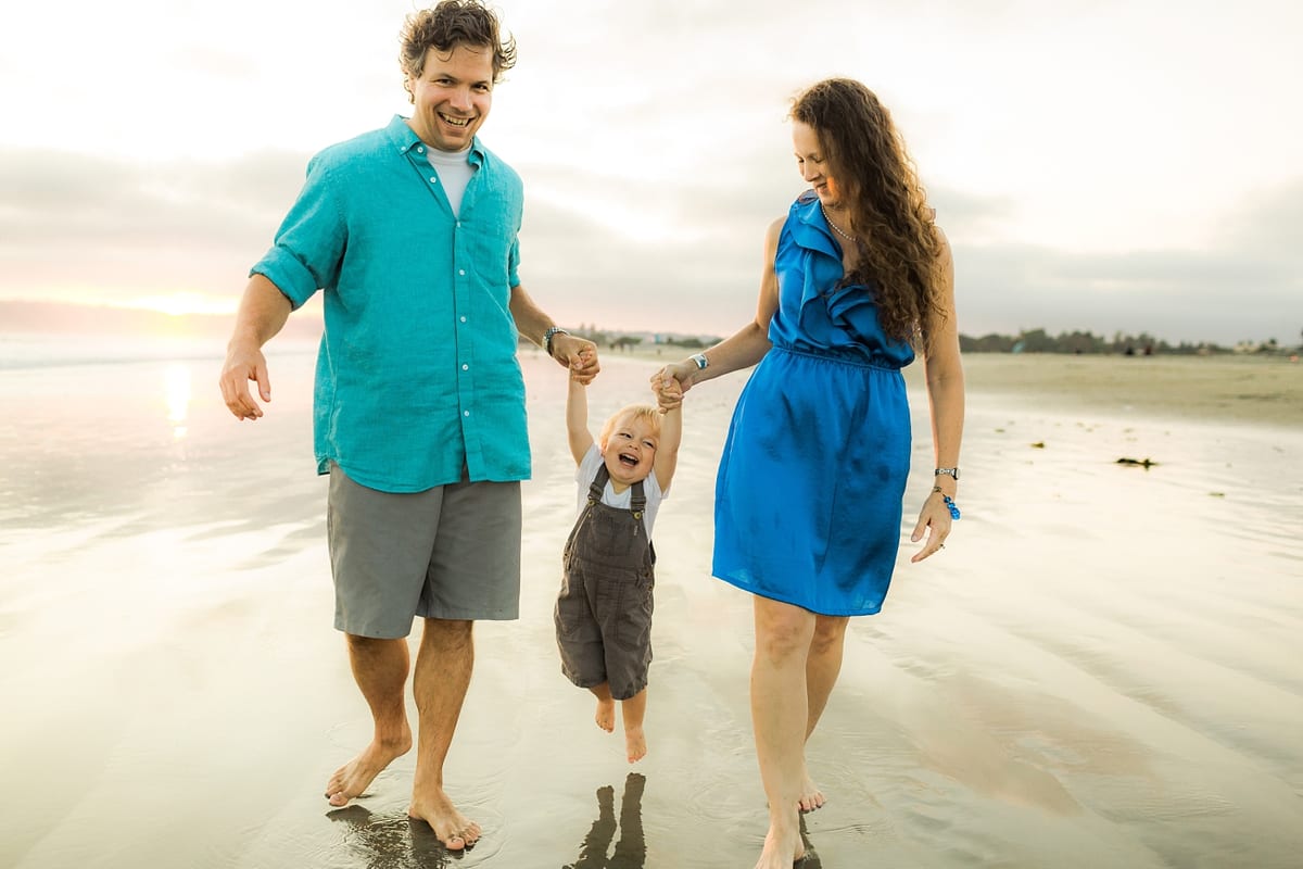 Parents swinging their toddler between them as they walk along the beach at sunset