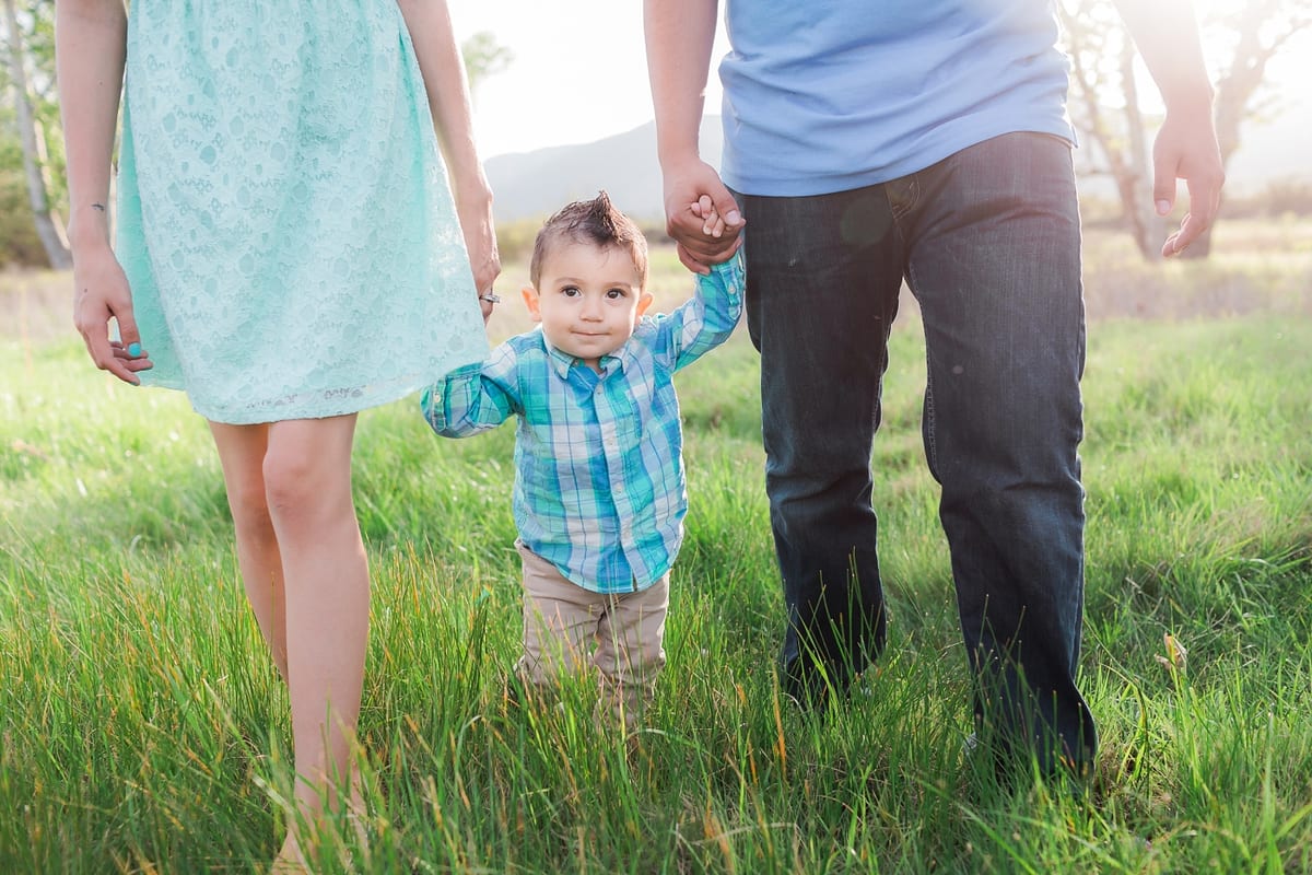 Toddler holding his parents' hands as he walks through the grass