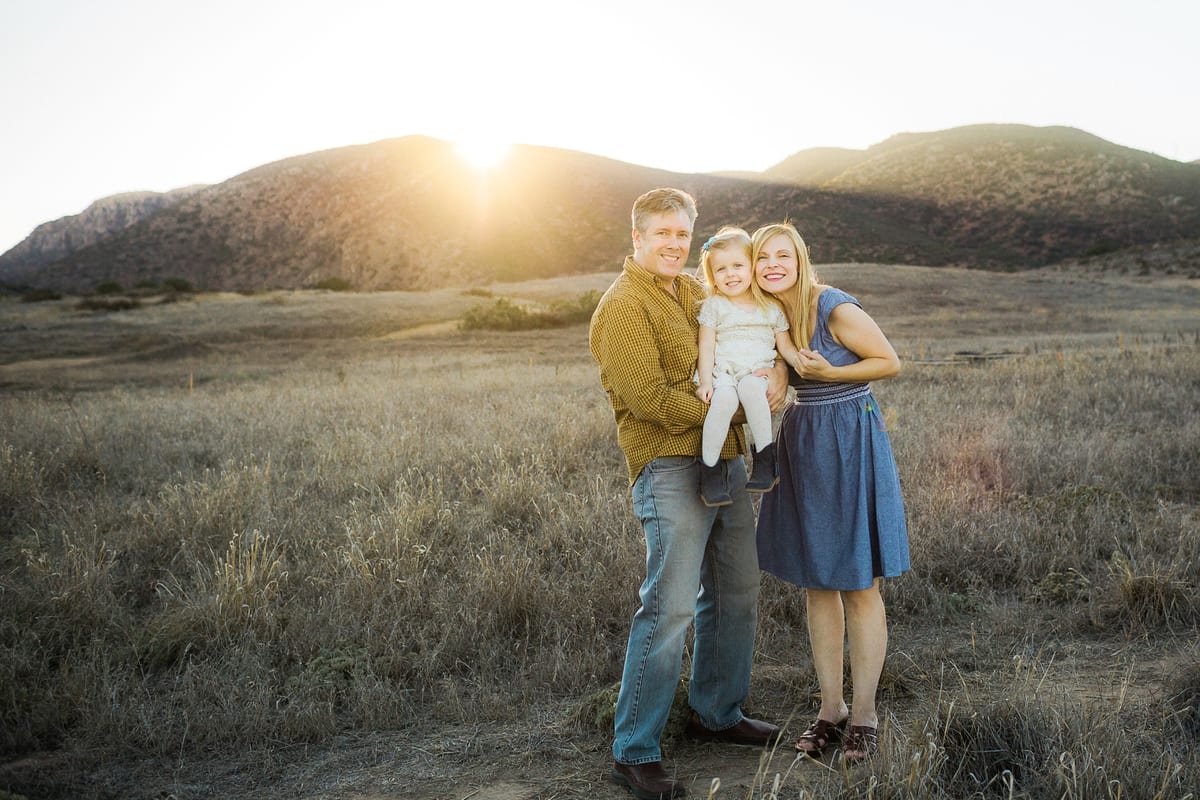 Parents holding their young daughter in a golden field at sunset with hills behind