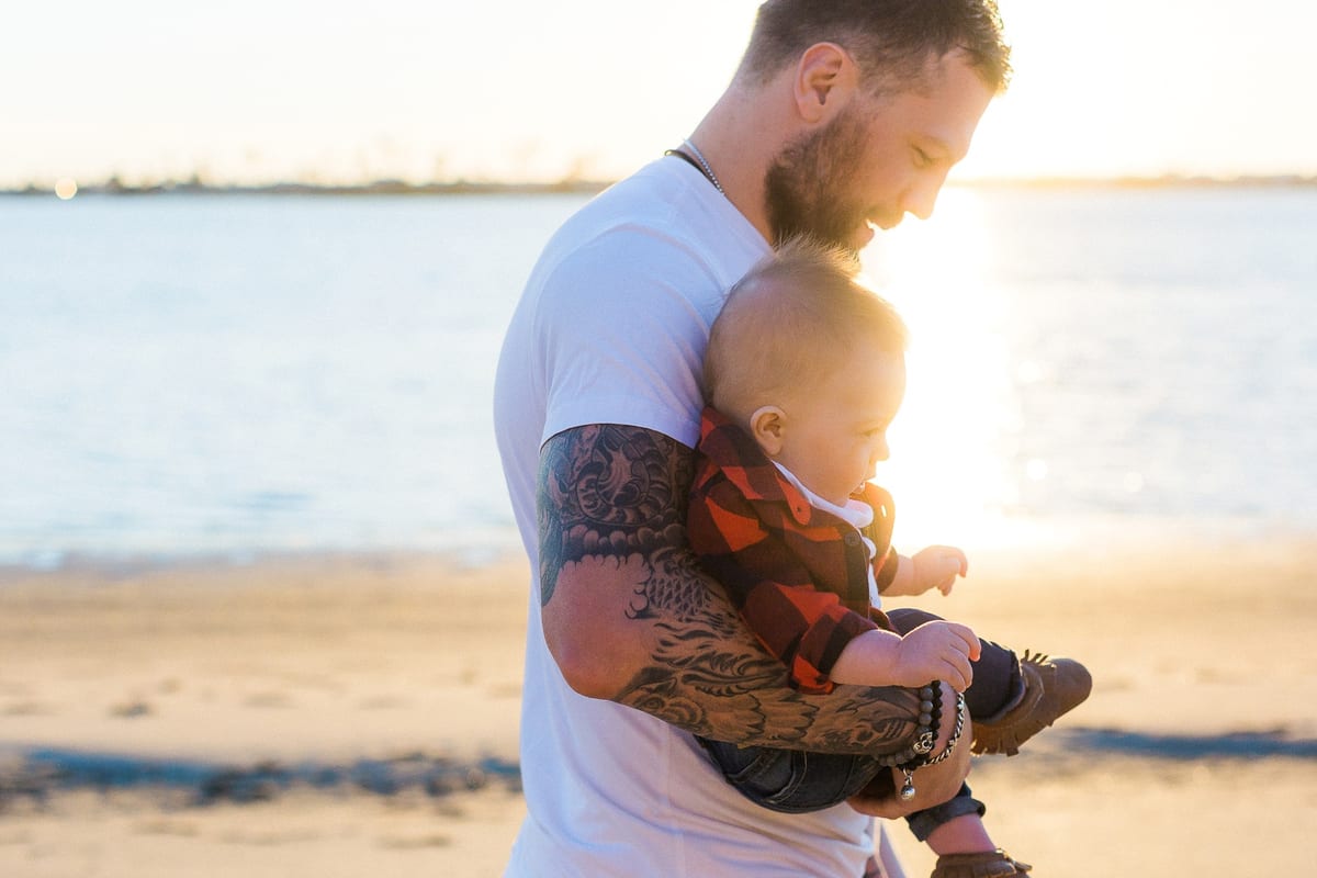 Tattooed father holding his baby son by the bay at sunset