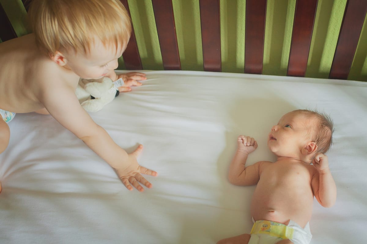 Toddler reaching out to touch his newborn sibling's hand in the crib