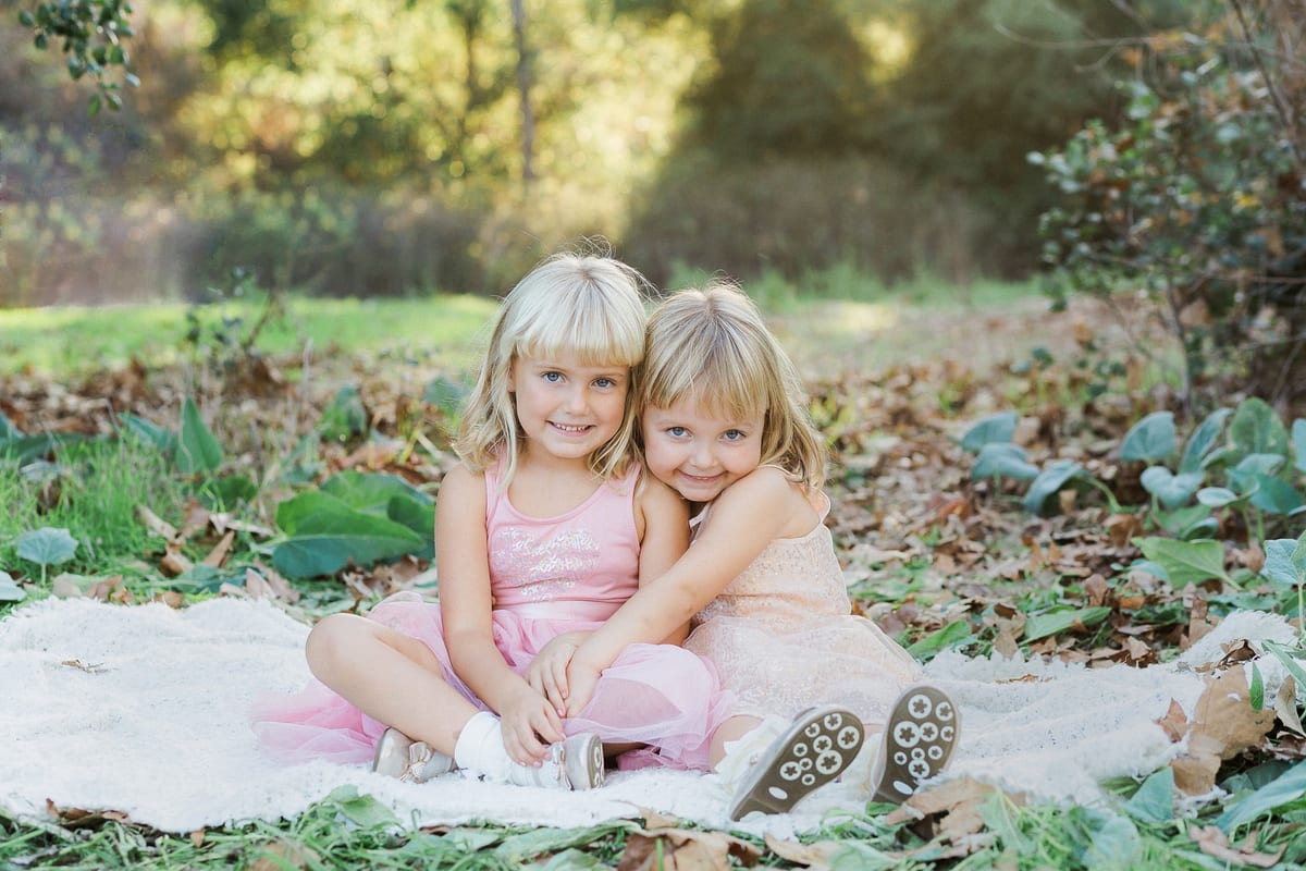 Two sisters in pink dresses sitting together on a blanket among autumn leaves