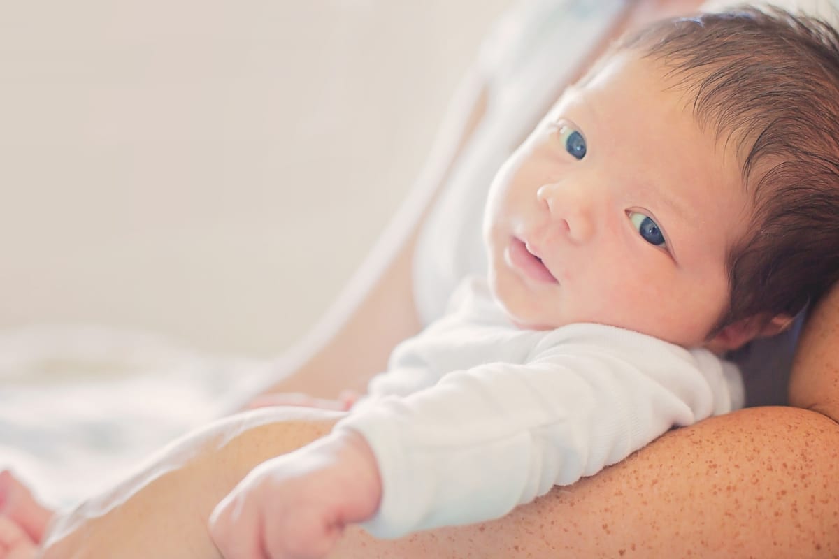 Newborn with dark hair gazing up while resting on a parent's arm