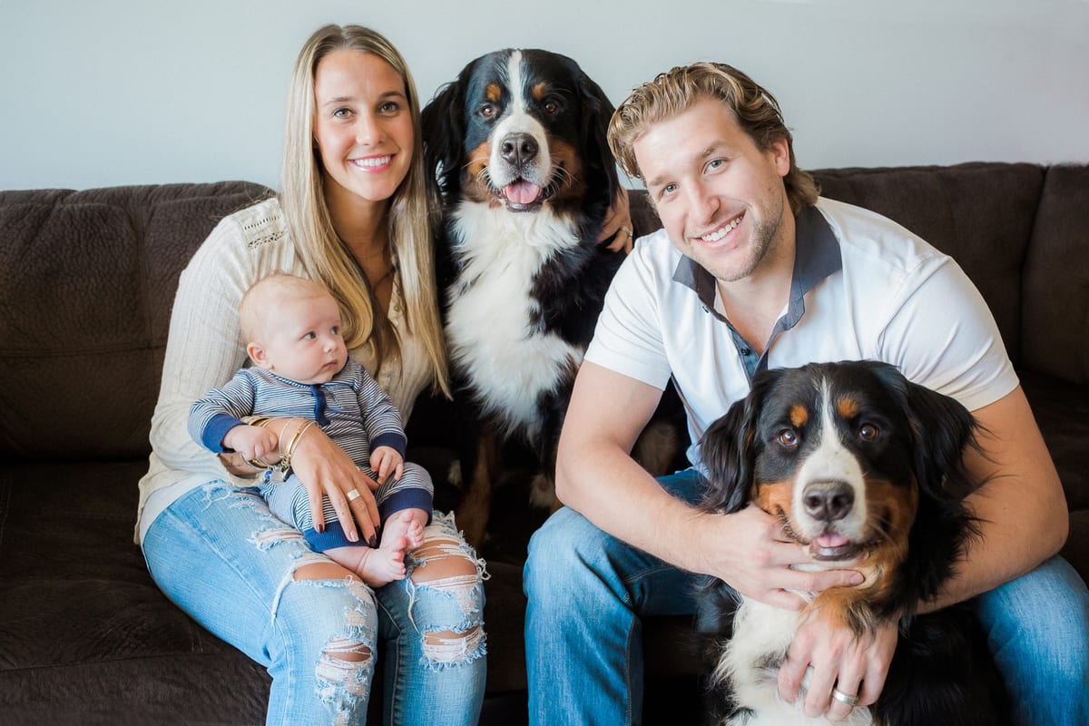 Parents smiling on the couch holding their baby with their two Bernese Mountain Dogs