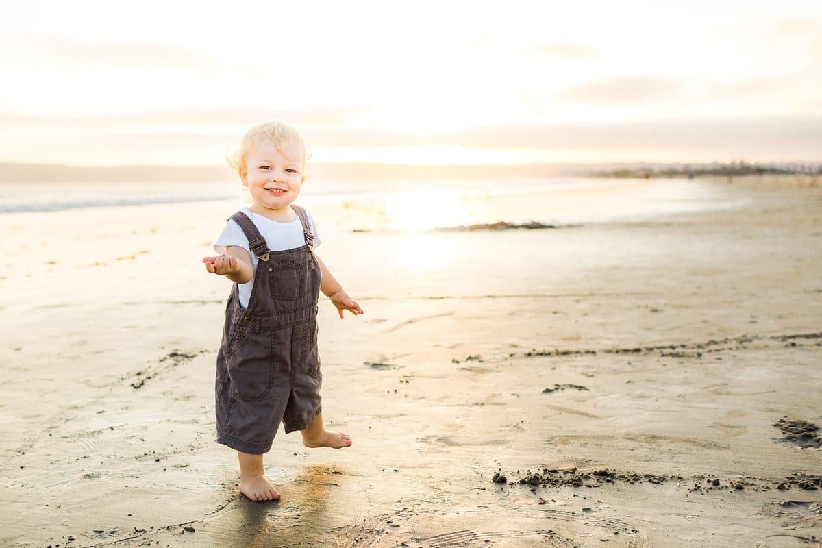Toddler in overalls walking barefoot on the beach at sunset