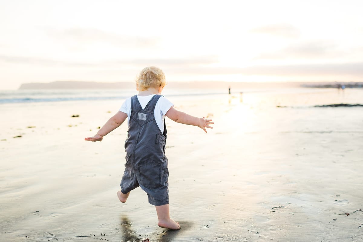 Toddler in overalls running along the wet sand toward the water
