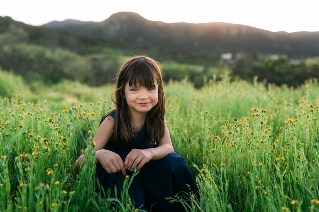 Smiling girl crouching in a field of yellow wildflowers below the mountains