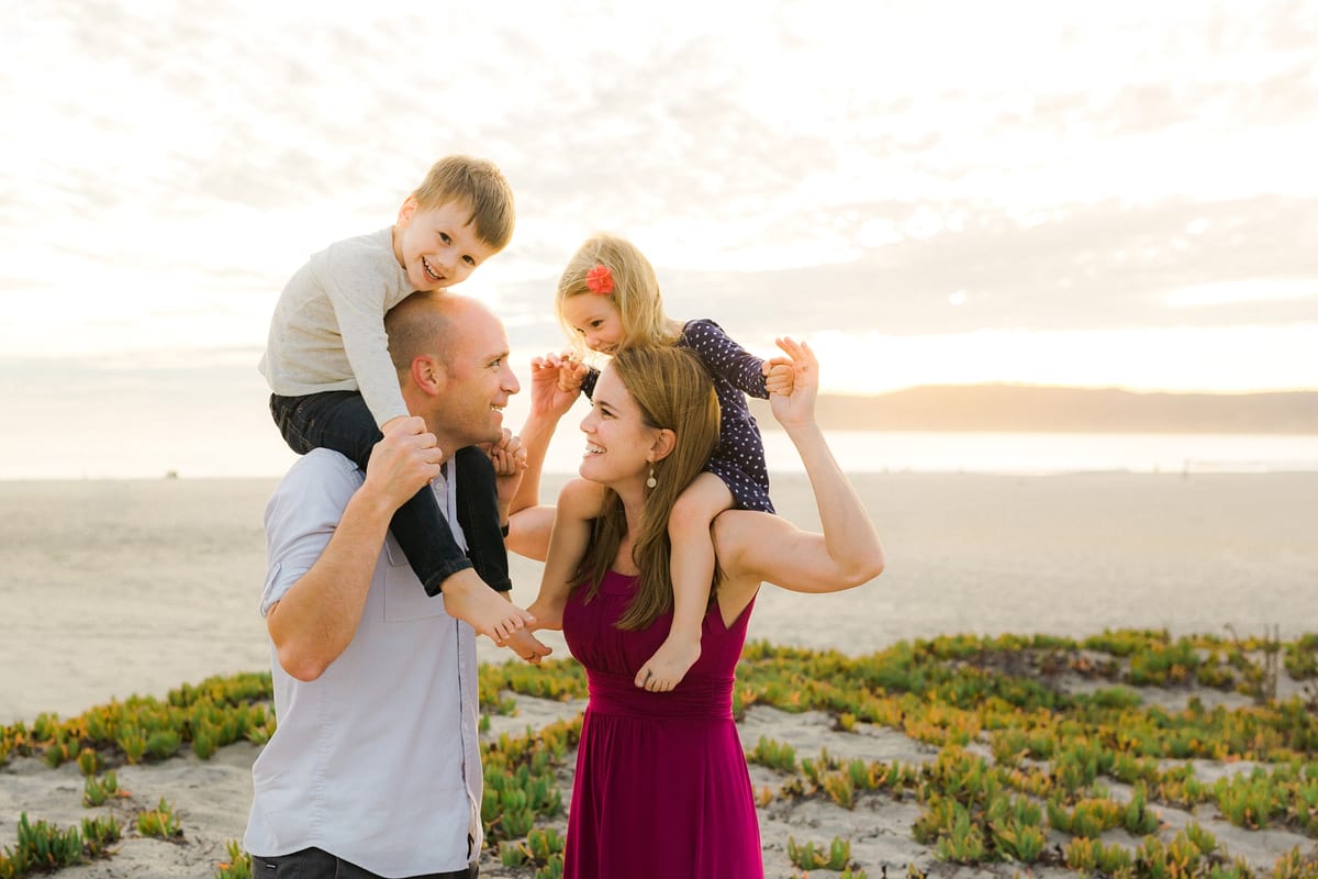 Children riding on their parents' shoulders on the beach at sunset